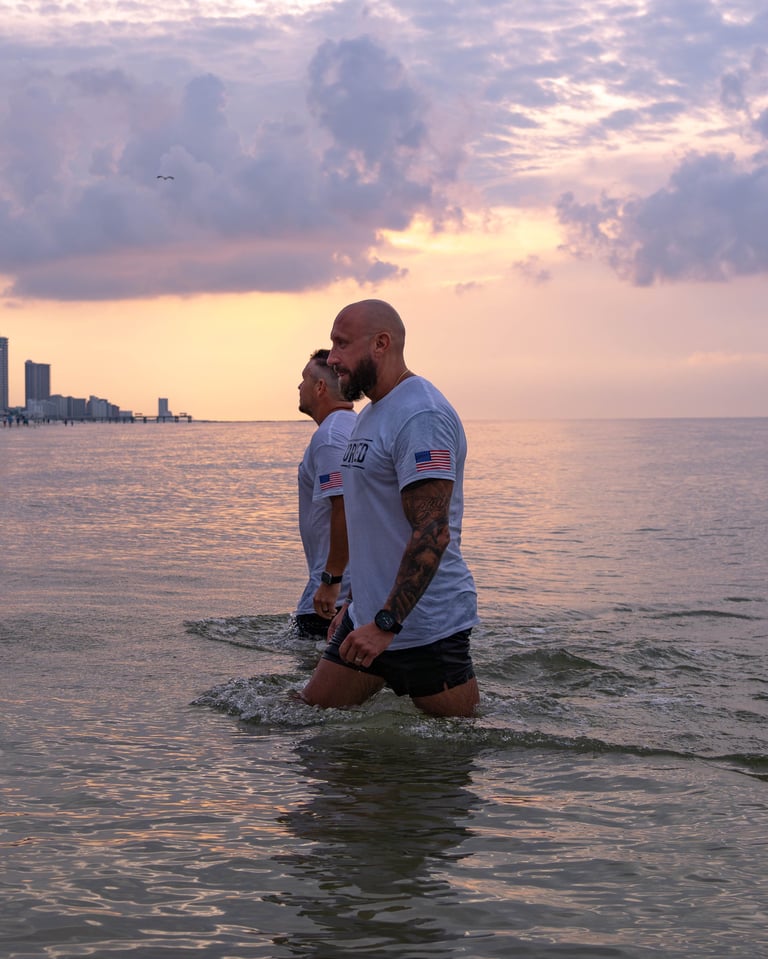 A man and woman embrace while standing in the ocean at sunset, with a city skyline visible in the background