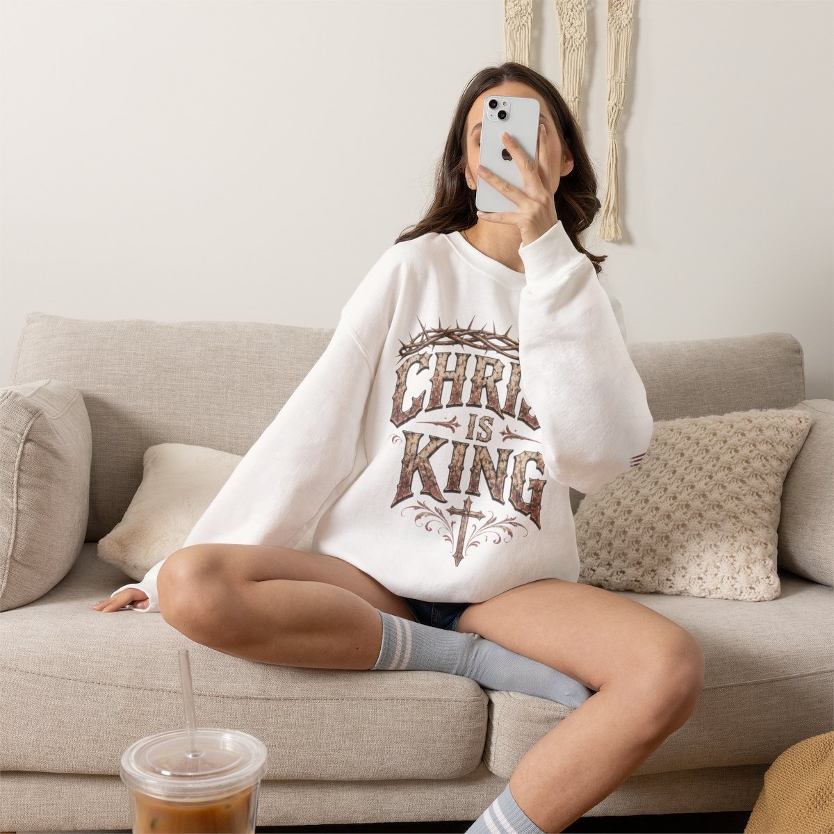 Person sitting on beige couch wearing oversized white graphic t-shirt and white socks, holding phone up to face