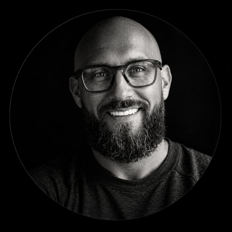Black and white portrait of a smiling man with glasses, beard, and shaved head wearing a dark t-shirt